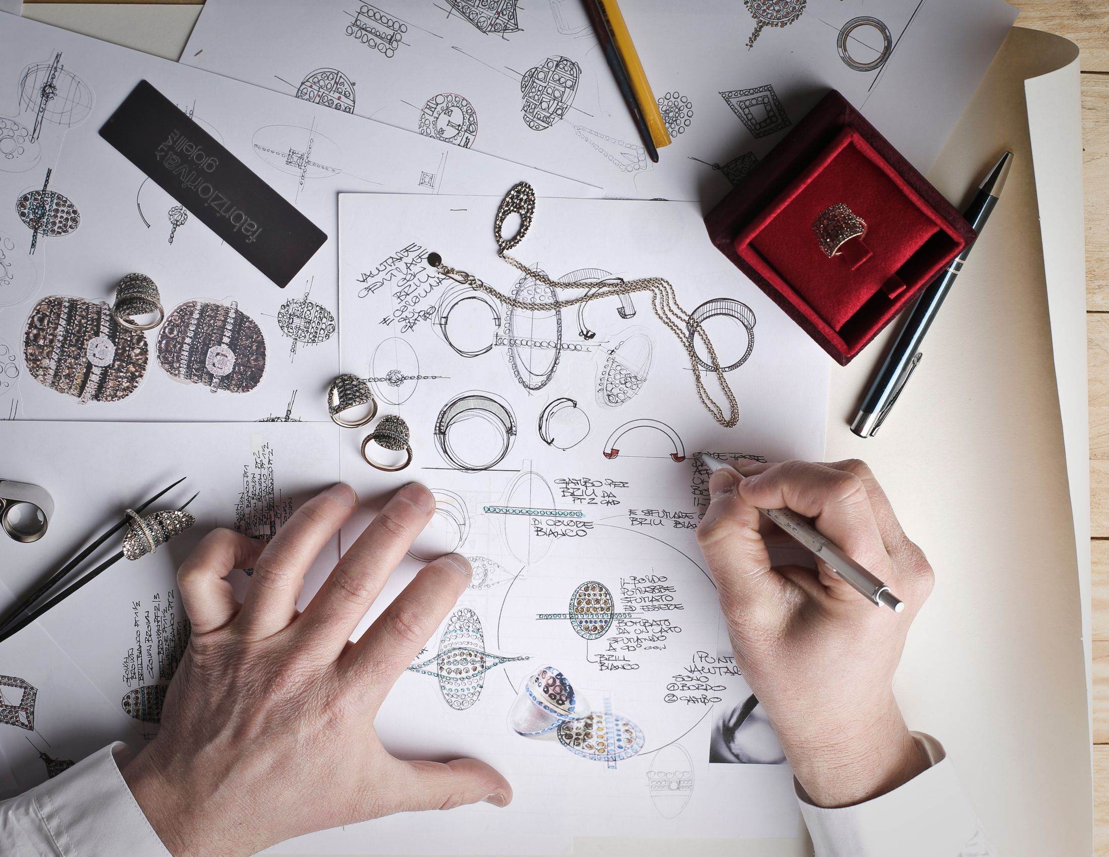 Hands sketching jewellery designs on paper beside a ring box and drawing tools