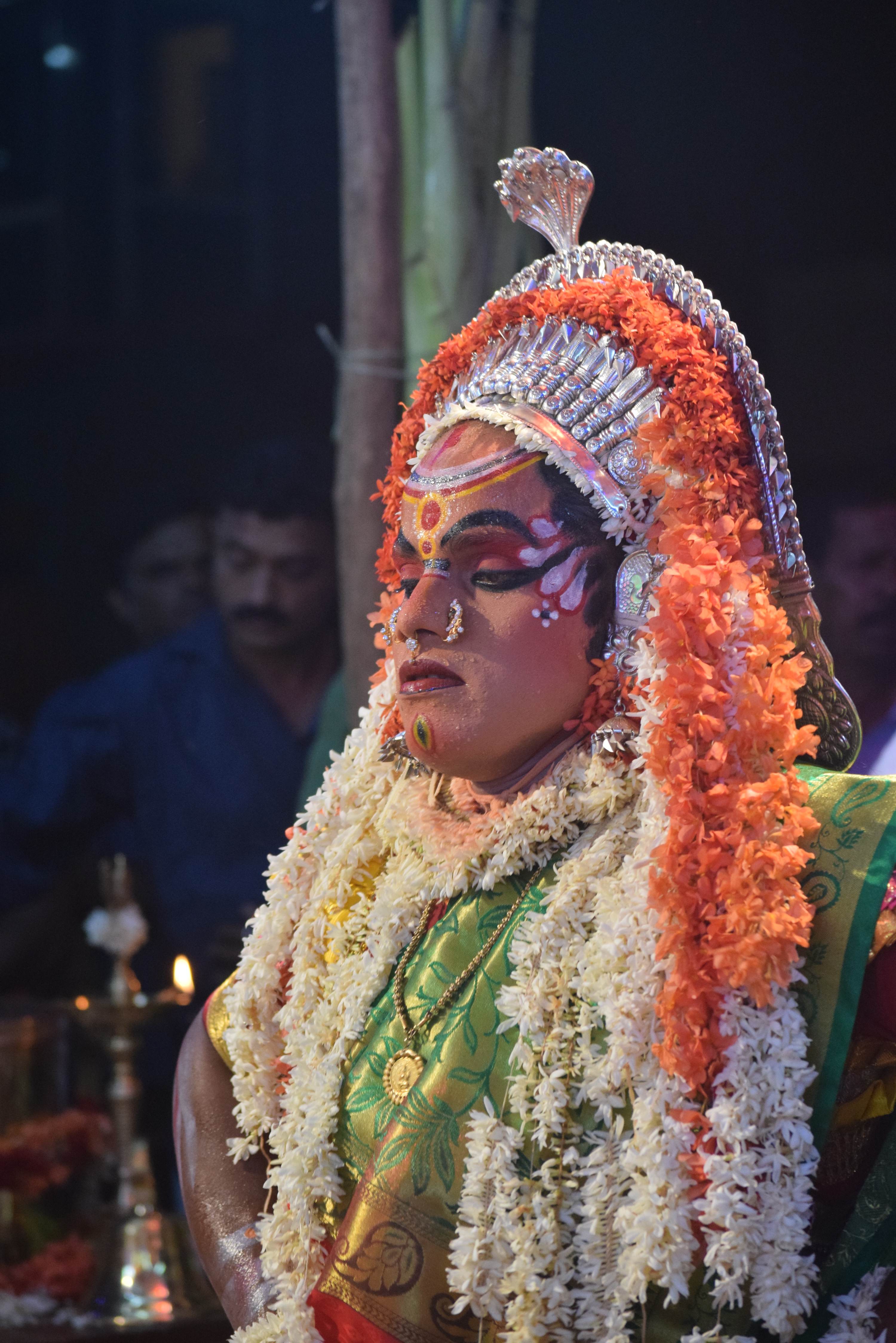A Kola performer in Mangaluru adorned with silver headgear and garlands of flowers during a night ceremony